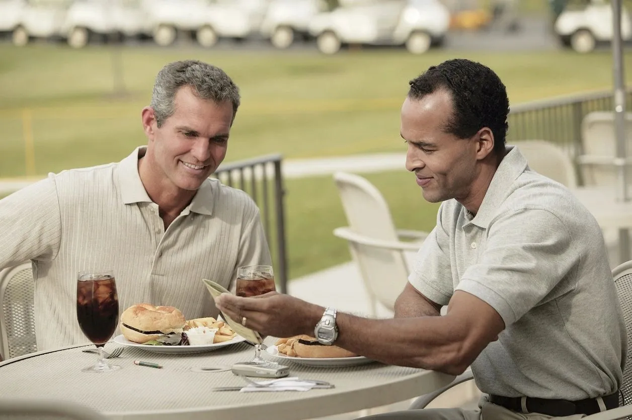 Two men sitting at a table eating sandwiches.